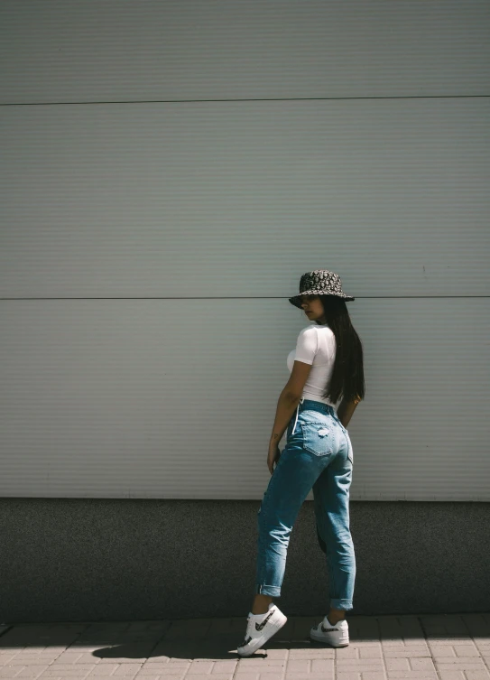 a girl walking against a wall wearing jeans and a hat