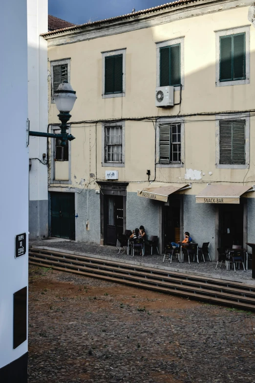 a group of people sitting outside of a building, by Nadir Afonso, old buildings, morning coffee, seen from a distance, grey