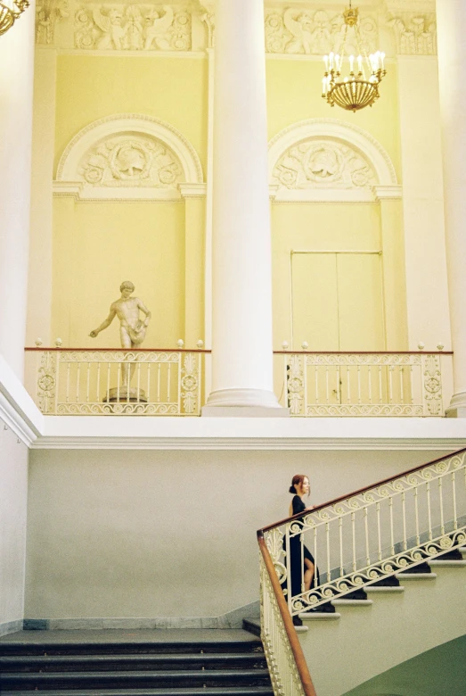 a person standing at the top of a set of stairs, inspired by Thomas Stothard, neoclassicism, pale yellow walls, museum sculpture, branching hallways, photo taken on fujifilm superia