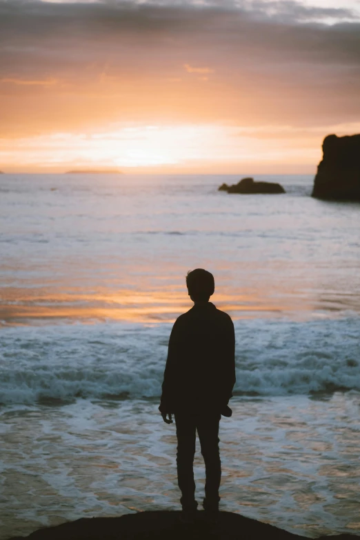 a man standing on top of a rock next to the ocean, by Jessie Algie, pexels contest winner, romanticism, teenage boy, back lit, abel tasman, faded glow