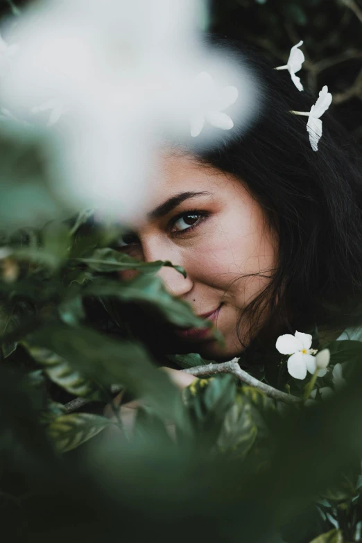 a woman with a flower in her hair, inspired by Elsa Bleda, trending on unsplash, lush foliage, hiding behind obstacles, smiling at the camera, zoomed in