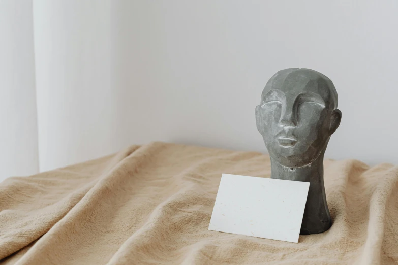 a sculpture sitting on top of a bed next to a blank card, trending on pexels, concrete art, portrait facial head, detailed product image, square face, decoration