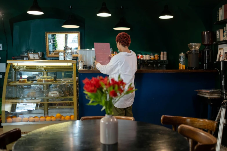 a woman standing in front of a counter at a bakery, by Liza Donnelly, pexels contest winner, private press, copper and deep teal mood, restaurant menu photo, flowers around, aussie baristas