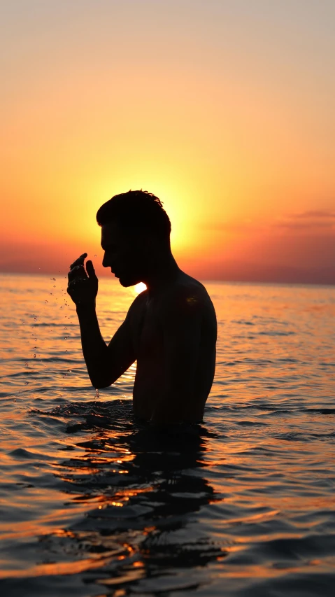 a man standing in the ocean at sunset, an album cover, by Alexis Grimou, pexels, drinking, lgbtq, solemn gesture, pondering