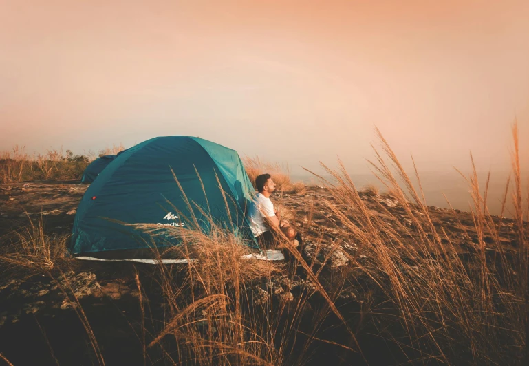 a man sitting in a field next to a blue tent, pexels contest winner, 🚿🗝📝, campsites, avatar image, looking onto the horizon