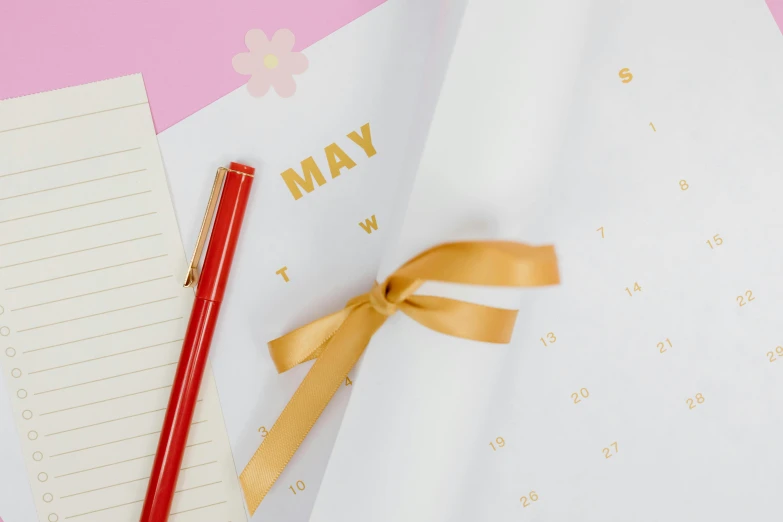 a red pen sitting on top of a piece of paper next to a calendar, by Julia Pishtar, trending on pexels, ornamental bow, may, pink and gold, max hay