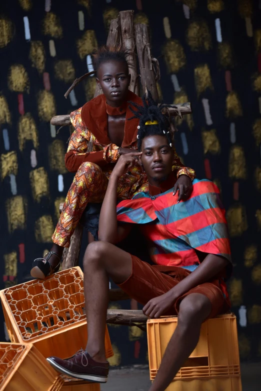 a couple of people sitting on top of crates, by Ingrida Kadaka, trending on pexels, afrofuturism, wearing a colorful men's suit, at a fashion shoot, a boy, sitting on intricate throne