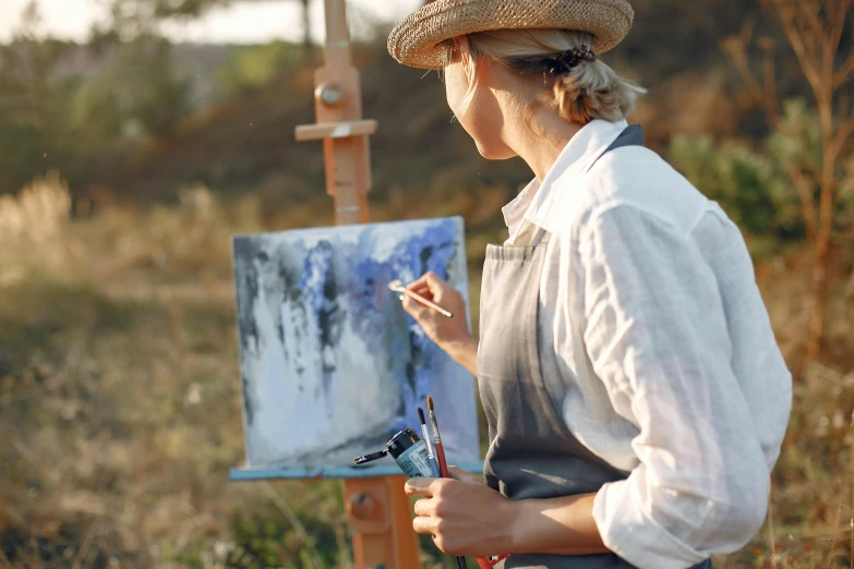 a woman in a straw hat painting on an easel, pexels contest winner, dominant wihte and blue colours, nature outside, holding a paintbrush, artist wearing overalls