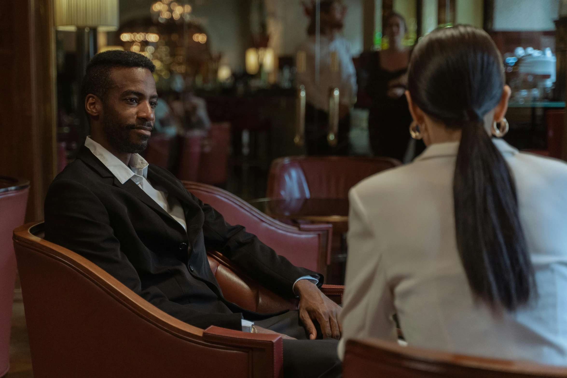 a man sitting in a chair talking to a woman, luxurious environment, a still of kowloon, riyahd cassiem, wearing a suits