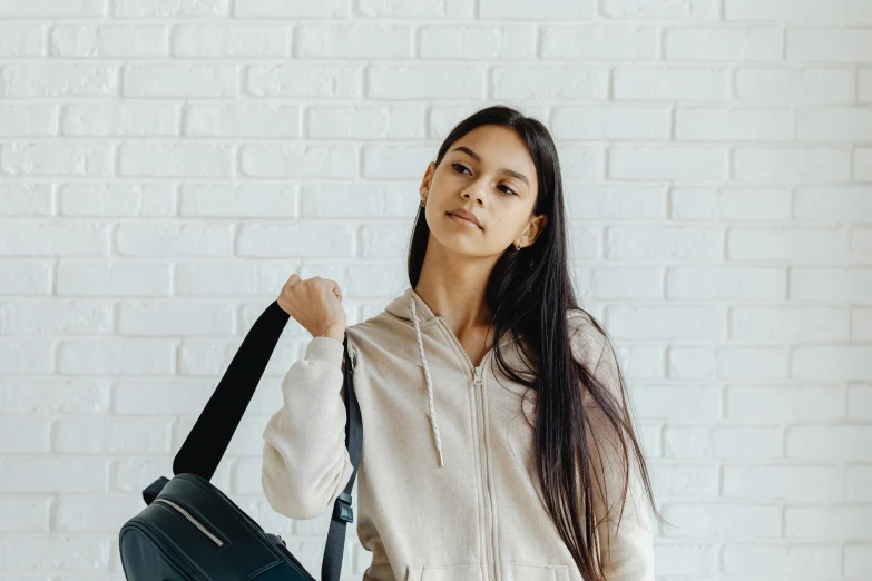 a woman holding a black bag in front of a white brick wall, trending on pexels, black haired girl wearing hoodie, avatar image, standing in class, sports clothing