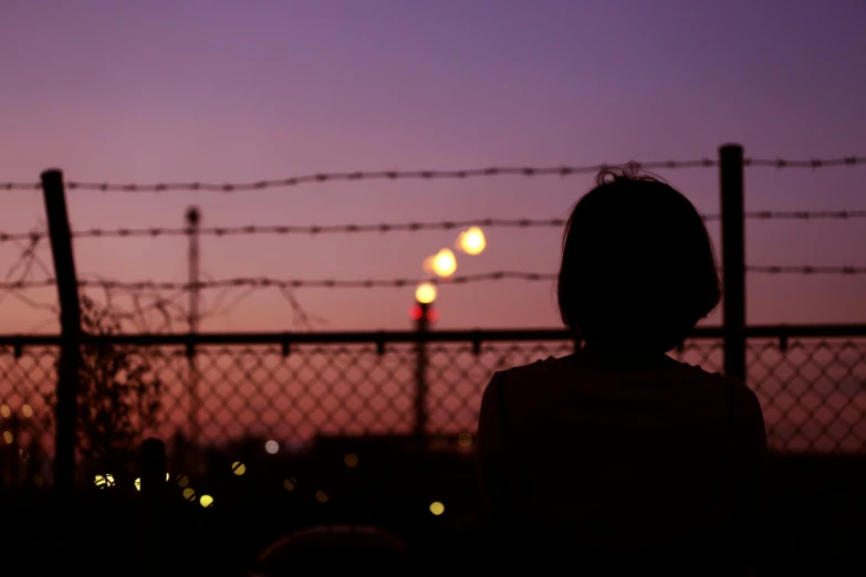 a silhouette of a person standing in front of a fence, inspired by Elsa Bleda, pexels contest winner, happening, girl sitting on a rooftop, lights in distance, violet sky, barbed wire