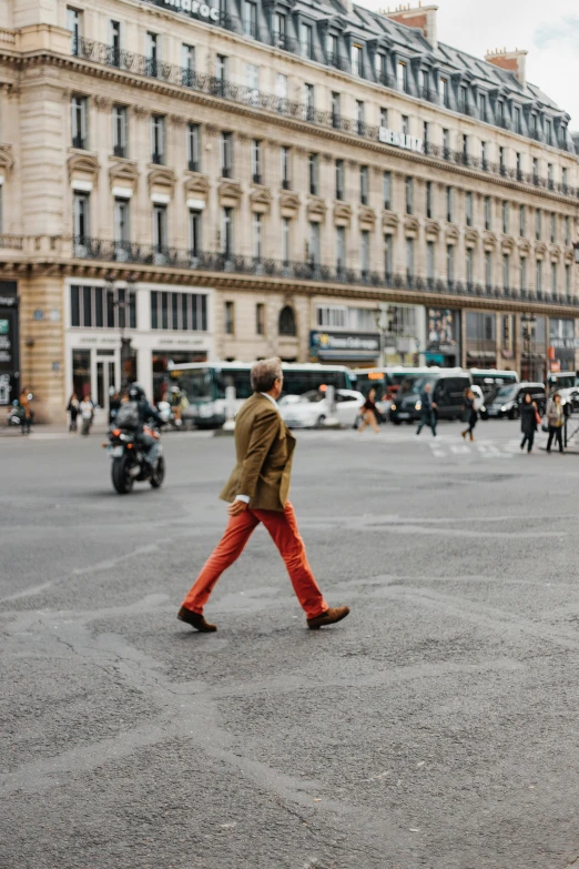 a man walking across a street next to tall buildings, by Raphaël Collin, trending on unsplash, red shirt brown pants, paris fashion week, orange gi, on a great neoclassical square