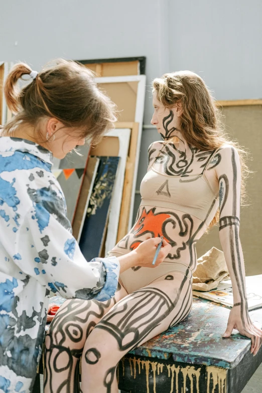 a woman sitting on top of a table next to another woman, inspired by artist, trending on pexels, process art, bodysuit, tribal paint, college, thin bodysuit