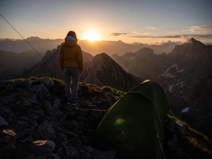 a person standing on top of a mountain next to a tent, sun down, in the dolomites, sun behind her, conde nast traveler photo