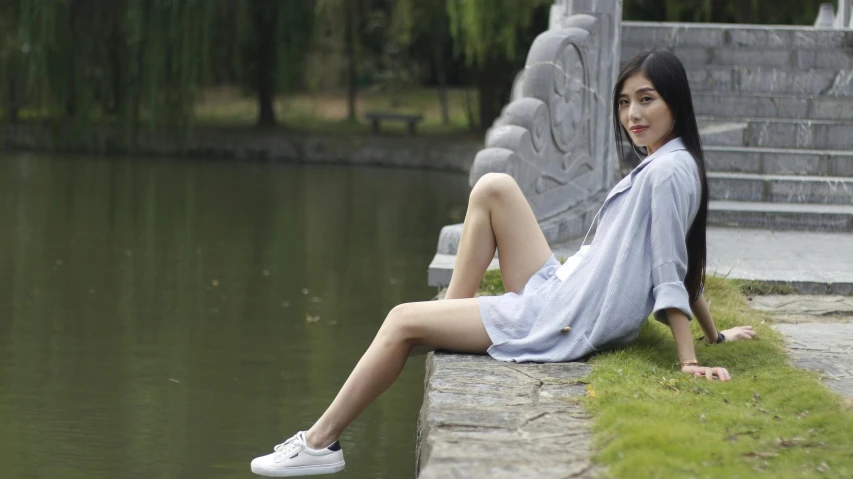 a woman sitting on a ledge next to a body of water, a portrait, inspired by Ma Yuanyu, pexels contest winner, white shoes, gemma chan beautiful girl, grey robe, wearing shorts and t shirt