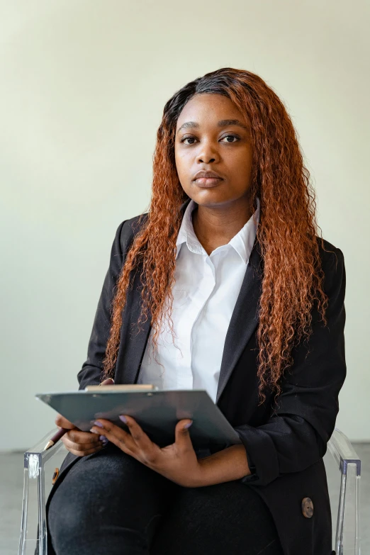 a woman sitting on a chair reading a book, with a business suit on, looking serious, dark-skinned, holding a clipboard