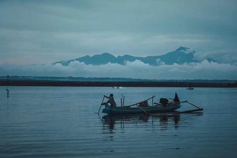 a man in a boat on a lake with a mountain in the background, by Emma Andijewska, pexels contest winner, sumatraism, shades of blue and grey, predawn, fish in the background, black volcano afar