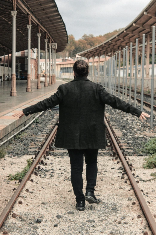 a man standing on a train track with his arms outstretched, an album cover, by Lucia Peka, unsplash, happening, wearing black overcoat, in spain, his arms are behind his back, train station