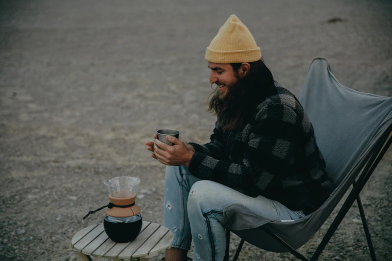 a man sitting in a chair with a cup of coffee, trending on unsplash, camping, with black beanie on head, aussie baristas, tall hat
