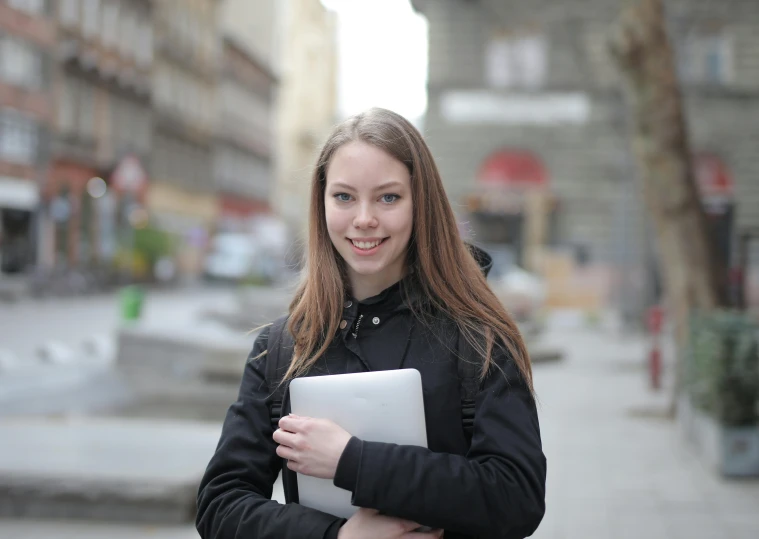 a woman in a black jacket holding a laptop, a portrait, pexels contest winner, portrait of white teenage girl, square, standing in street, avatar image