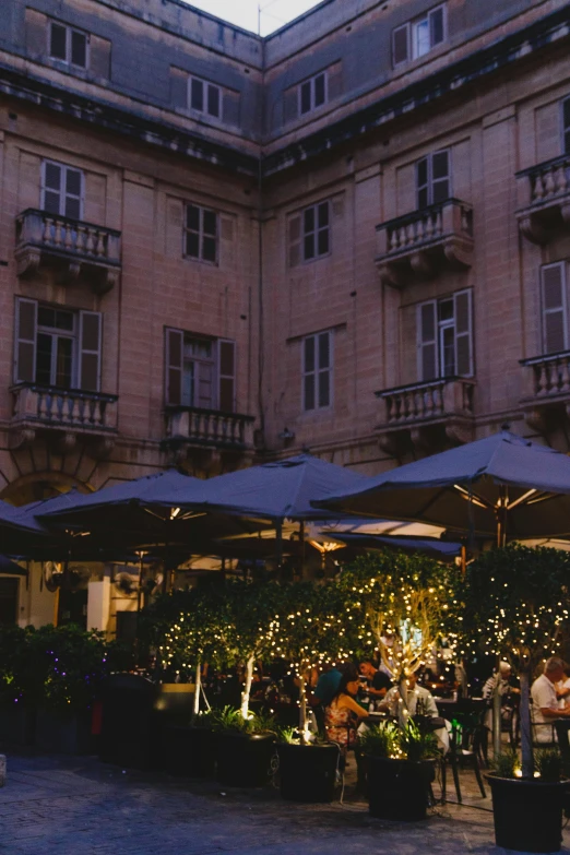 a group of people sitting under umbrellas in front of a building, in the evening, lush, al fresco, surrounding the city