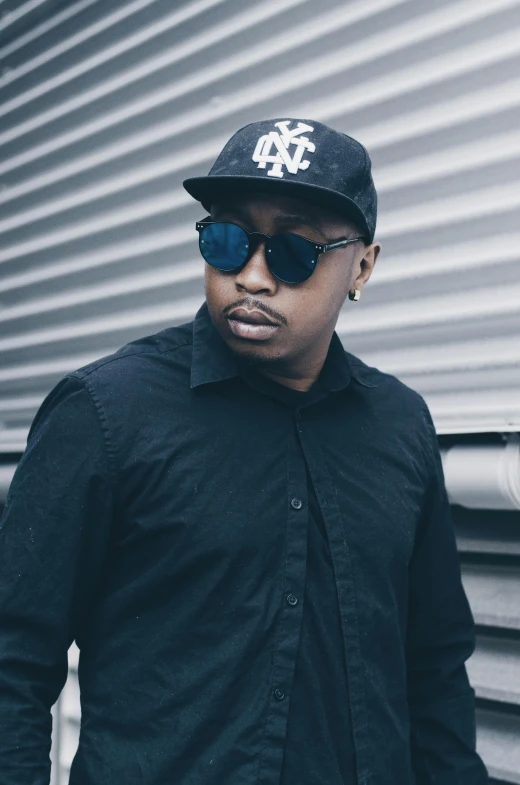 a man standing in front of a metal wall, an album cover, inspired by Zhu Da, trending on unsplash, big floppa, his frank blue eyes, wearing sunglasses and a cap, looking to the side off camera