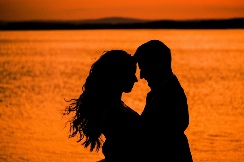 a couple standing next to each other in front of a body of water, by Matt Cavotta, pexels contest winner, romanticism, orange sun set, profile image, stylized silhouette, headshot