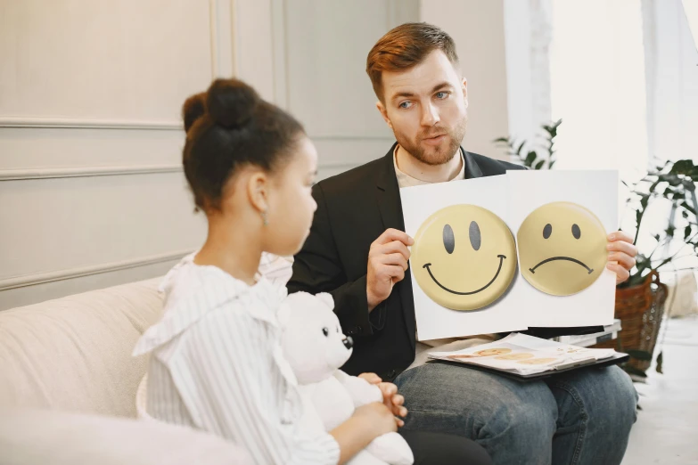 a man and a woman sitting on a couch holding a piece of paper with smiley faces drawn on it, a photo, by Julia Pishtar, trending on pexels, analytical art, clinically depressed, woman holding sign, brown smiling eyes, professional picture