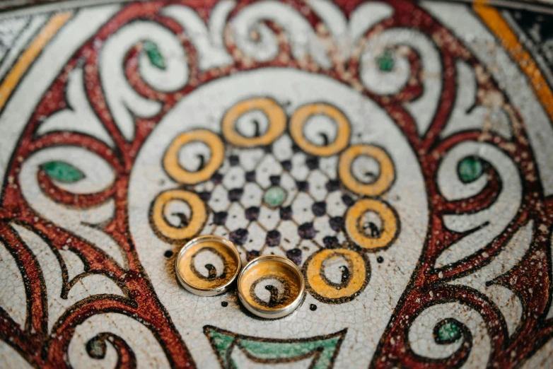 a couple of wedding rings sitting on top of a table, a mosaic, inspired by Taddeo Gaddi, trending on unsplash, cloisonnism, ocher details, high details photo, curled perspective, ancient stone tiling