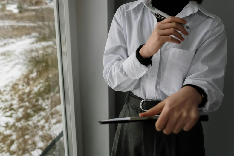 a woman standing in front of a window holding a cell phone, a picture, trending on pexels, visual art, in white turtleneck shirt, holding a clipboard, wearing a shirt with a tie, hand holding a knife