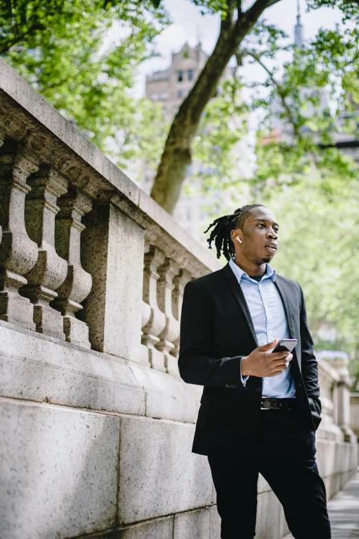 a man in a suit standing next to a stone wall, an album cover, inspired by Richard Wright, pexels contest winner, renaissance, looking at his phone, in a city park, man is with black skin, young man with medium - length