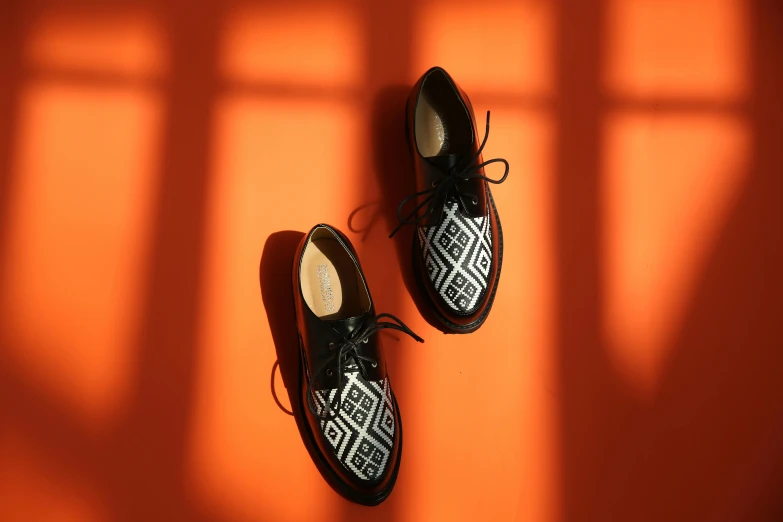 a pair of black shoes sitting on top of an orange wall, inspired by Mi Fu, trending on pexels, op art, with backdrop of natural light, aztec, intricate image, creepers