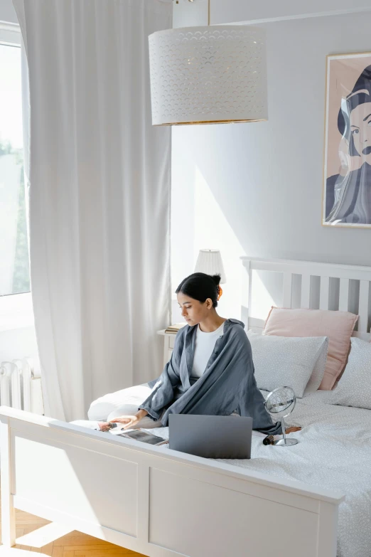 a woman sitting on a bed using a laptop, inspired by Li Di, grey robe, airbnb, home office, slightly minimal