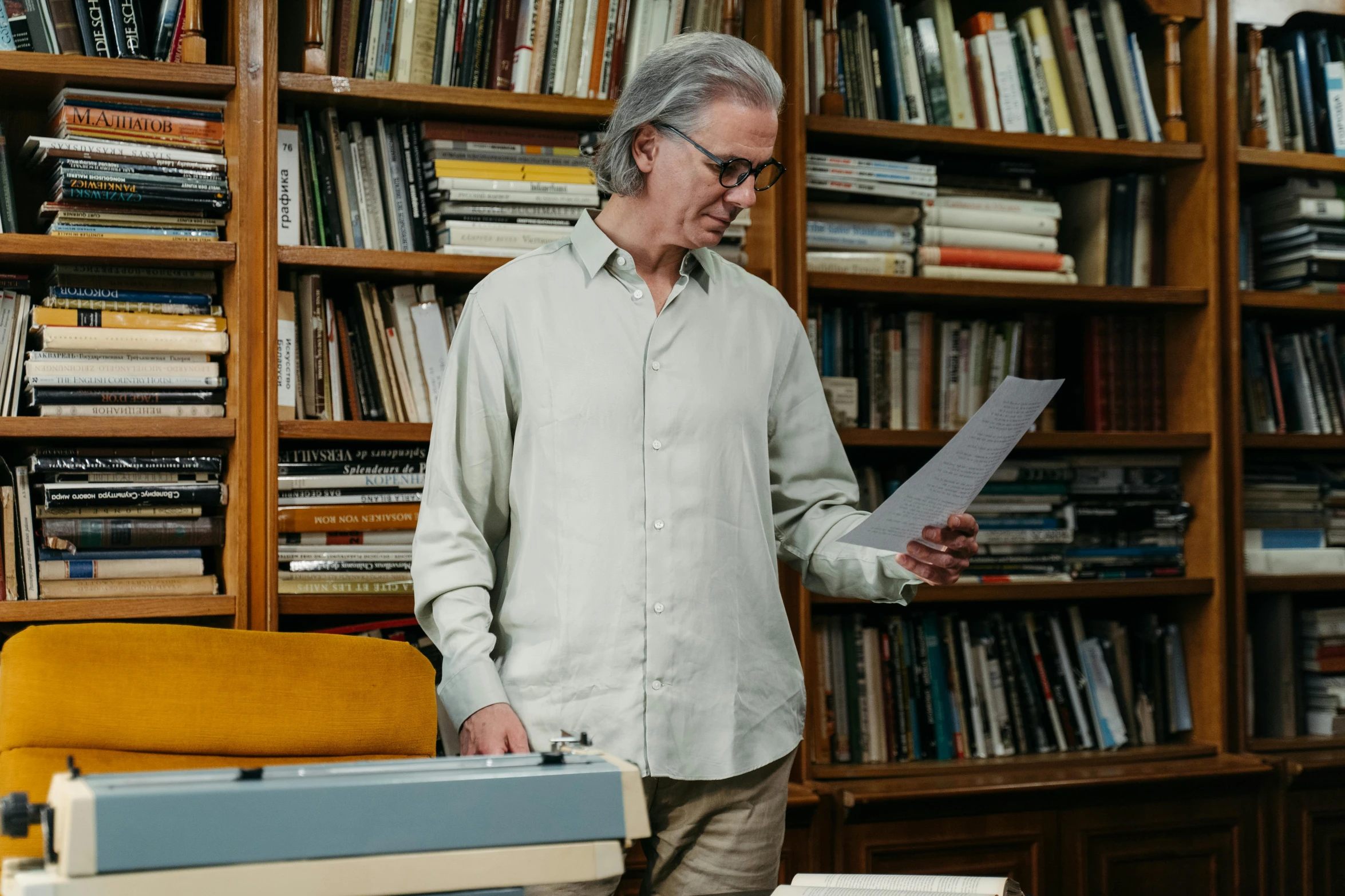 a man standing in front of a bookshelf holding a piece of paper, by Bertram Brooker, pexels, private press, a silver haired mad, wooden desks with books, wes anderson), programming