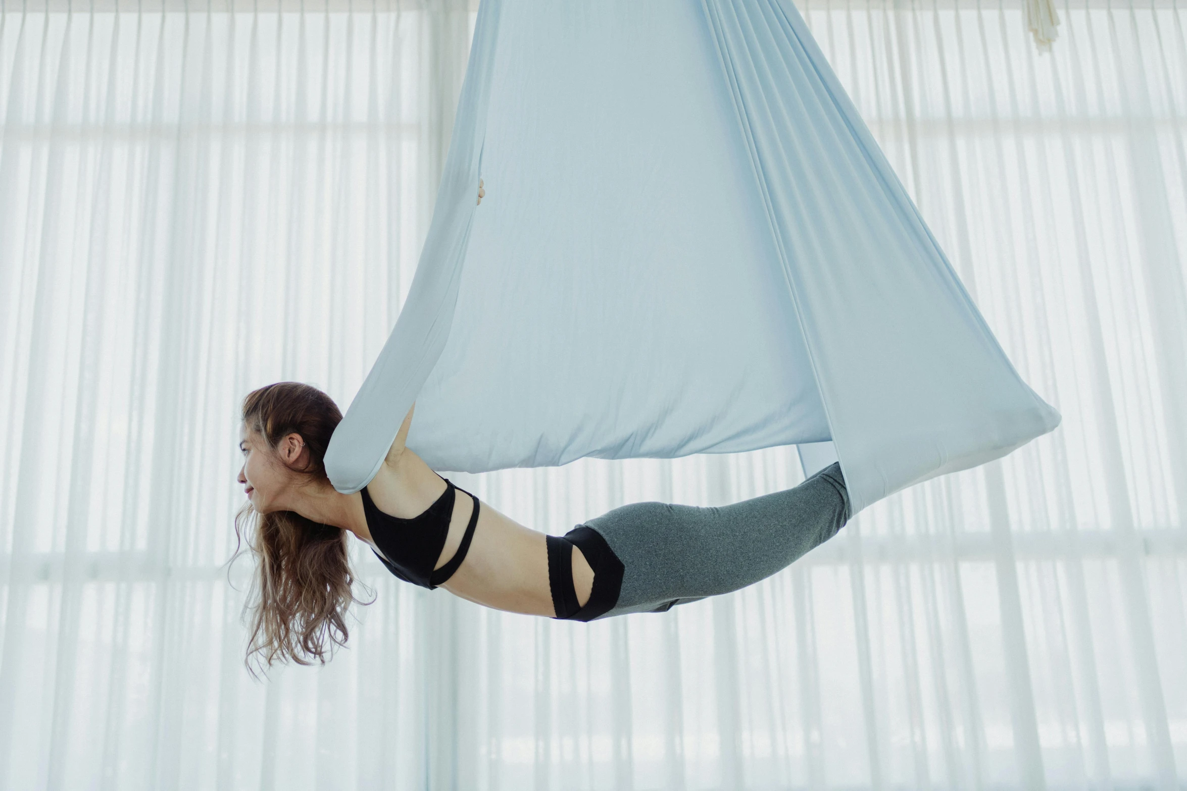 a woman hanging upside down on a hammock, by Emma Andijewska, arabesque, silks, dingy gym, sky blue, medium-shot