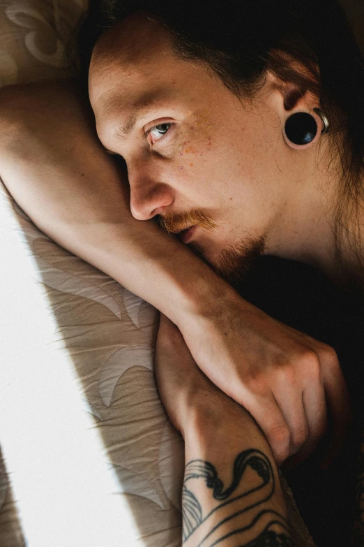 a man laying on a bed with a tattoo on his arm, by Jan Tengnagel, trending on pexels, visual art, backlit ears, androgynous face, pondering, looking out