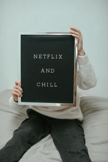 a person sitting on a bed holding a sign that says netflix and chill, by Everett Warner, trending on pexels, serial art, chalkboard, foil, chell, cold
