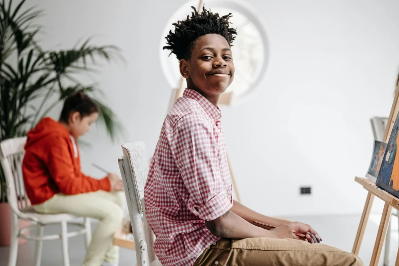 a man sitting on a chair in front of a painting, by Lily Delissa Joseph, pexels contest winner, black teenage boy, checkered spiked hair, happy kid, wearing a linen shirt