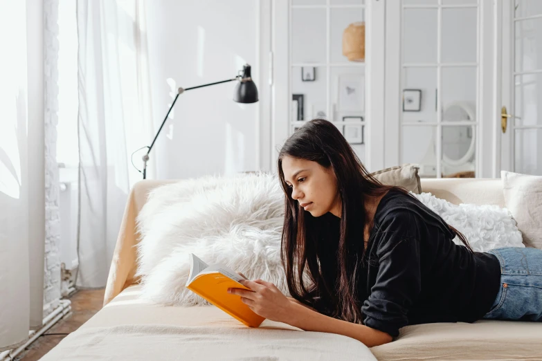 a woman laying on a bed reading a book, a cartoon, pexels contest winner, sitting on couch, reading engineering book, black haired girl wearing hoodie, lady using yellow dress