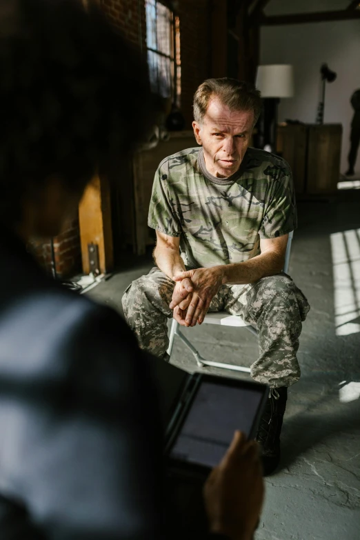a man sitting in a room with a laptop, rugged soldier, sad prisoner holding ipad, teaching, military-grade