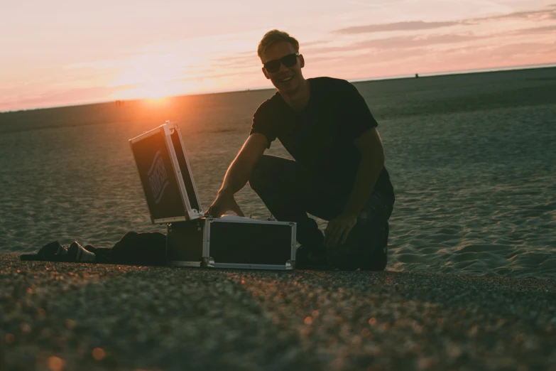 a man sitting on the ground next to a suitcase, a picture, unsplash, happening, mellotron, standing on the beach at sunset, posing for camera, radio box