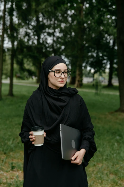 a woman in a black hijab holding a cup of coffee, inspired by Maryam Hashemi, pexels contest winner, hurufiyya, walking at the park, wearing square glasses, holding notebook, looking serious