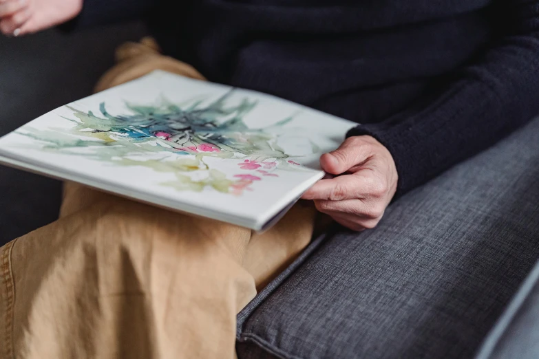 a person sitting on a couch holding a book, a detailed painting, inspired by Jenny Saville, featured on unsplash, botanical herbarium paper, angled shot, watercolours, pressed flowers