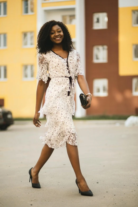 a woman walking across a parking lot next to a tall building, pexels contest winner, happening, dressed in a frilly ((lace)), black young woman, happy fashion model, flowery dress