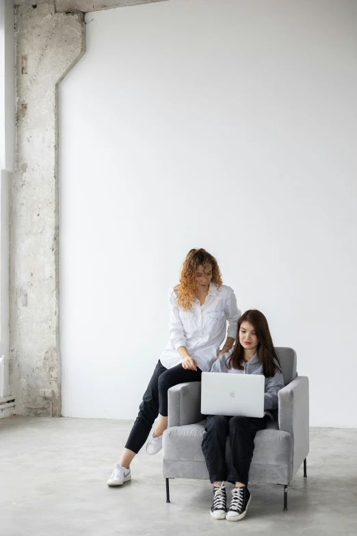 two women are sitting on gray couches looking at their laptops