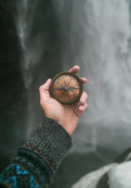 hand holding small wooden compass near large waterfall