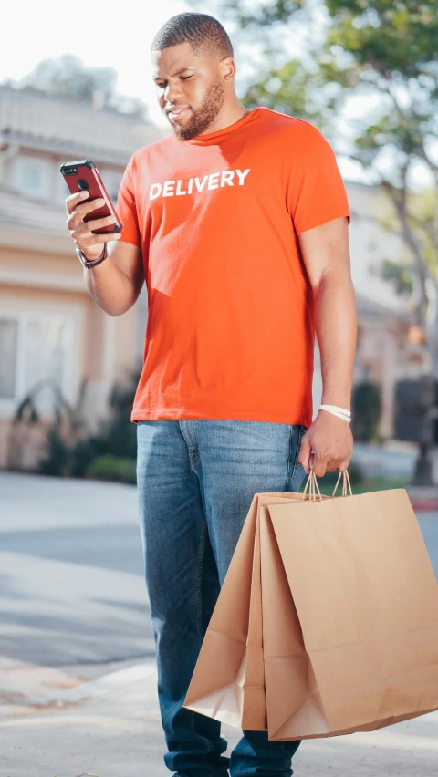 a man stands on a curb holding two bags and looking at his cell phone