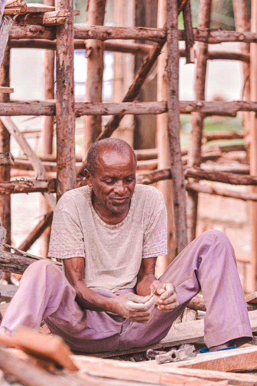 an elderly man sits on the ground in front of a house made out of wood