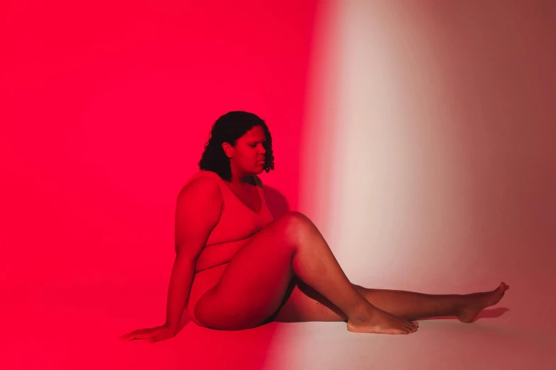 a woman in a red bodysuit sitting in front of a pink background