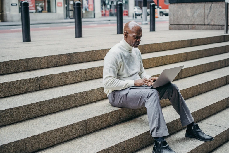 a man in a white sweater and grey pants sits on some steps while using his laptop